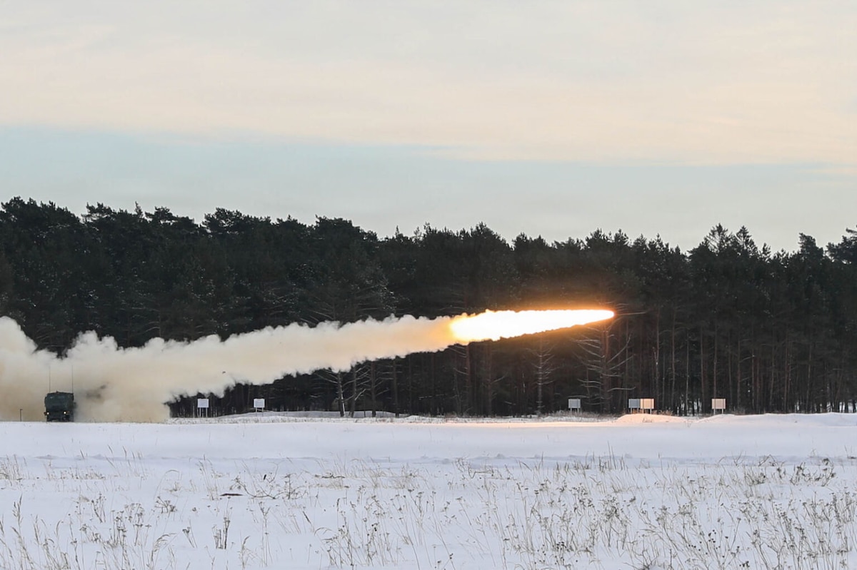 A rocket glides through the air over snow-covered ground after being fired from a launching system. A line of trees can be seen in the background.