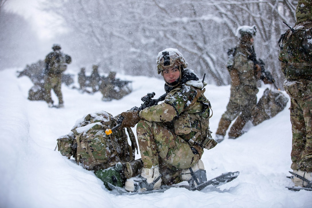 Snow falls as soldiers in winter gear kneel while holding weapons in a snowy field surrounded by trees.