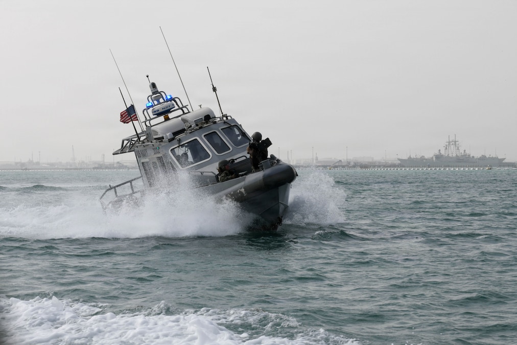 A boat with blue sirens and an American flag on a top sails to the right while transiting a body of water on a gloomy day.