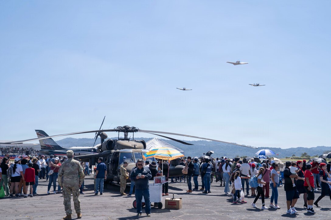 A crowd surrounds a military helicopter and airplanes fly in the background