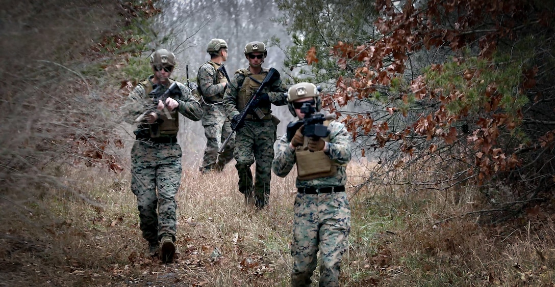 4 Marines walking through the woods with foliage with 4 rifles that are equipped with the organic counter small UAS systems.