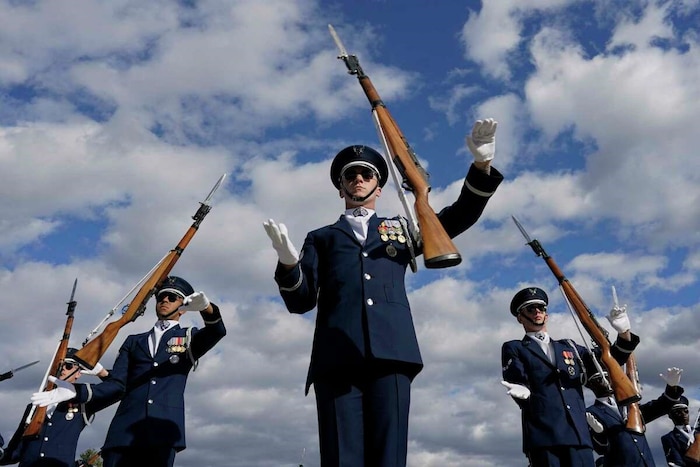 A U.S. Air Force Honor Guardsman stands alongside other drill team members. His rifle is spinning in the air directly above him. He wears a formal dark blue ceremonial uniform with white gloves.