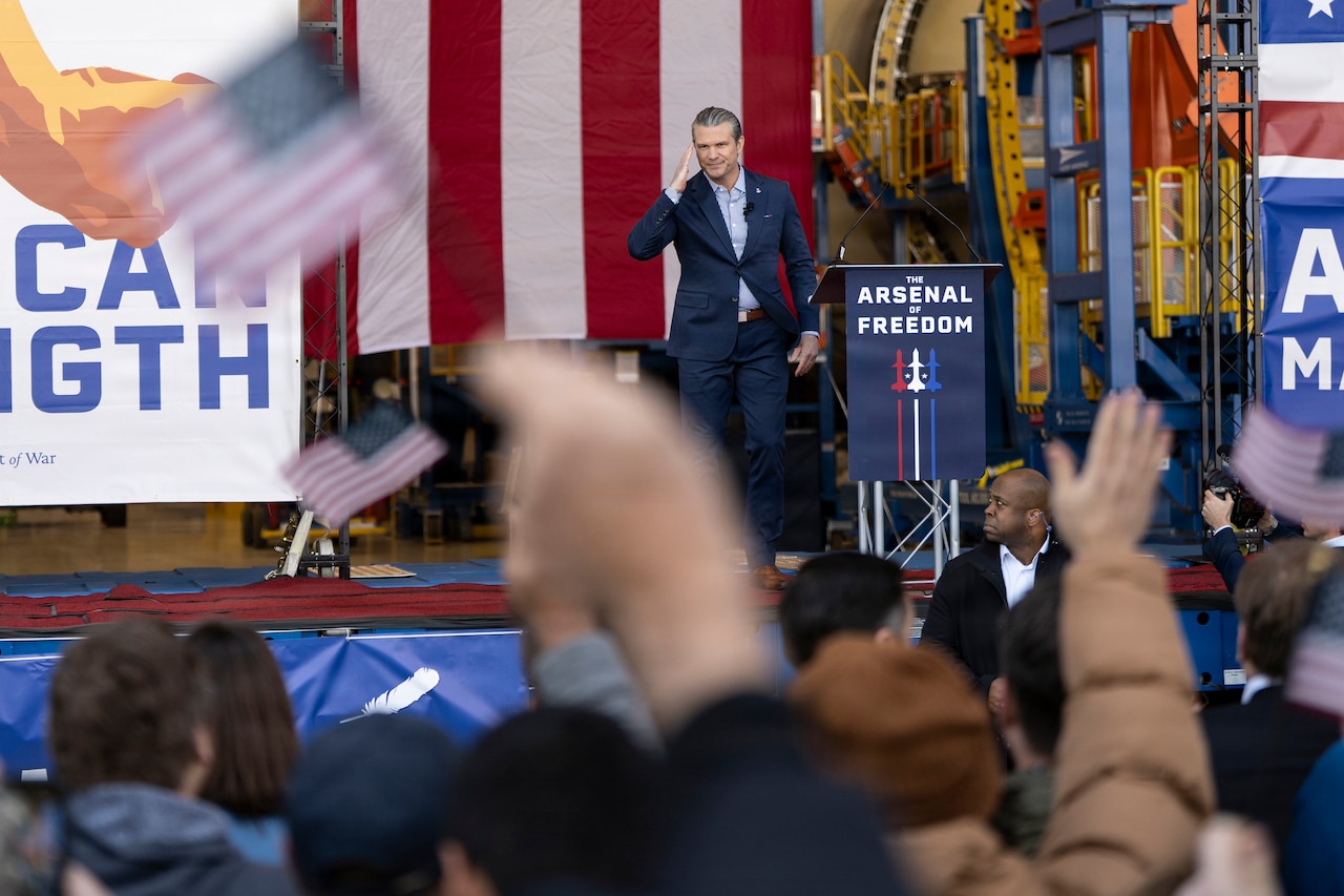 A man in a business suit stands on a stage saluting a crowd of people with his right hand; the lectern next to him reads, “Arsenal of Freedom.” In the foreground are people with their arms up, some waving small American flags.