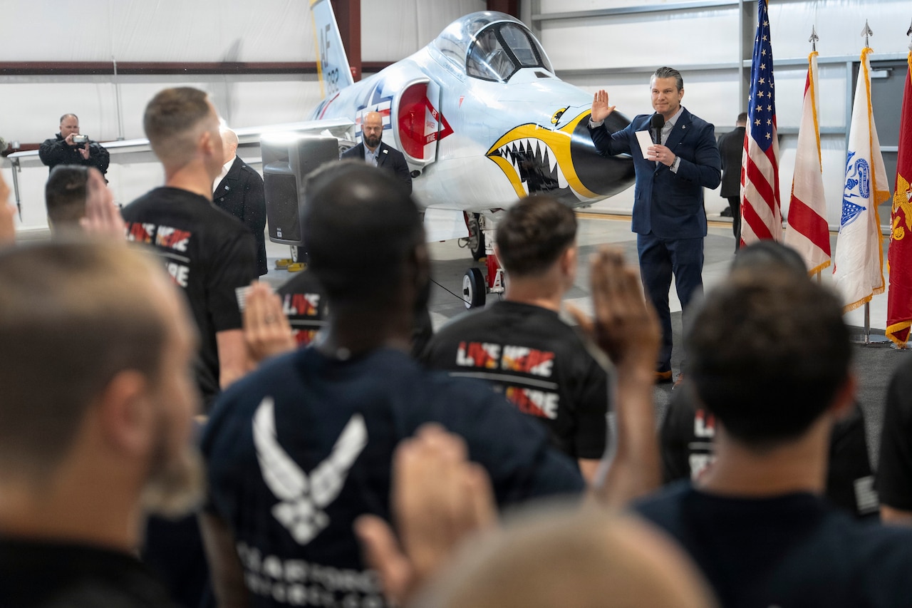 A man in a business suit stands inside an aircraft hangar with his right hand raised while holding a piece of paper in his opposite hand; behind him are a military jet, an American flag and three other flags. In front of the man, a group of people stands with their backs to the camera and their right hands raised.