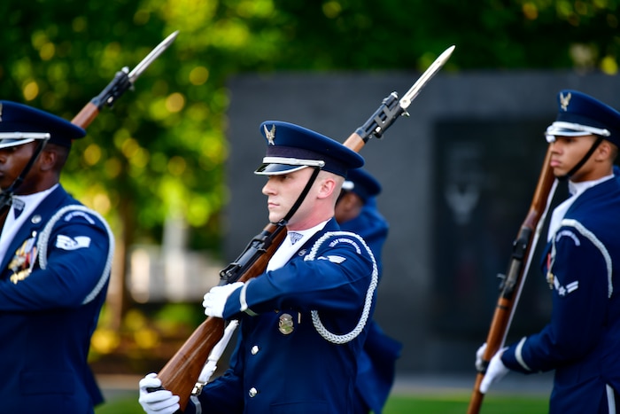 A U.S. Air Force Honor Guardsman stands alongside other drill team members. His rifle rests on his shoulder. He wears a formal dark blue ceremonial uniform with white gloves.