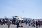 A crowd surrounds a military helicopter and airplanes fly in the background
