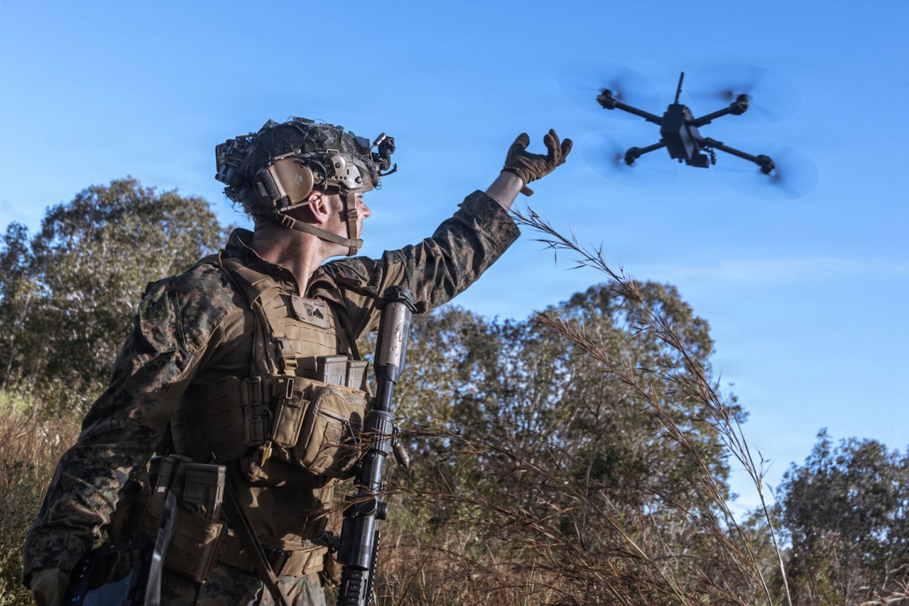 A man in a camouflage military uniform stands in thick brush while holding his hand in the air as a drone flies away.