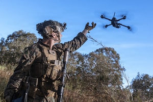 A man in a camouflage military uniform stands in thick brush while holding his hand in the air as a drone flies away.