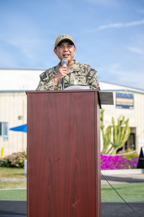 A man in a camouflage military uniform speaks into a microphone while standing behind a lectern. There is a large building in the background.