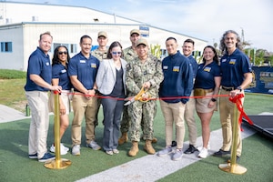 About a dozen people in athletic attire and camouflage military uniforms stand in front of a ribbon as a man in a camouflage military uniform uses large scissors to cut the ribbon. There is a large building in the background.
