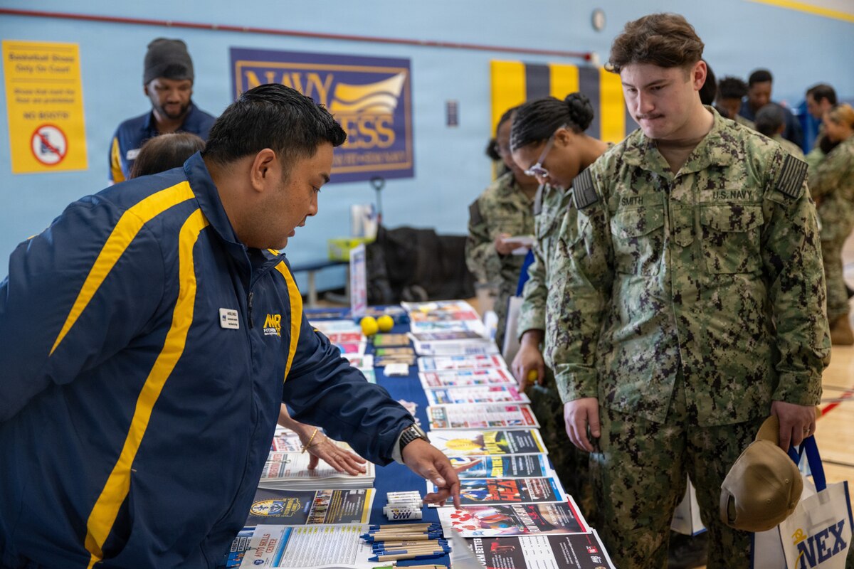 A man in athletic attire points to pamphlets on a table as another man in a camouflage military uniform looks down at the table. There are several people in camouflage military uniforms standing in the background.