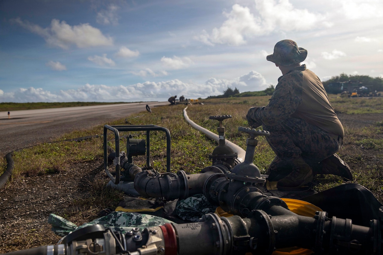 A man in a camouflage military uniform looks off into the distance while turning on the faucet of a long water hose in a grassy terrain on the side of a road under a blue sky with clouds; other military personnel are at the far end of the hose.