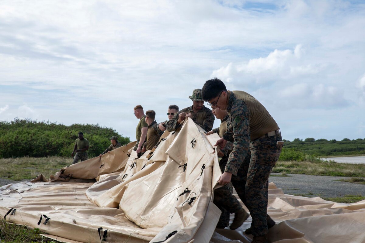 Nearly a dozen people in camouflage military uniforms lift a large beige tarp in a grassy terrain on a cloudy day.