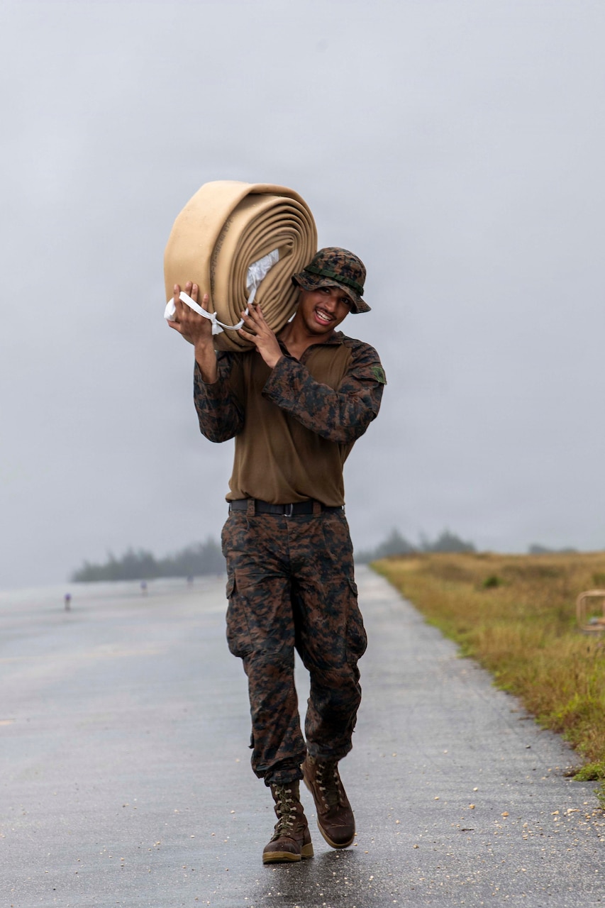 A man wearing a camouflage military uniform smiles while walking on a paved road and holding a large, wrapped water hose on his shoulder.