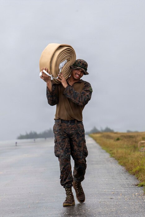 A man wearing a camouflage military uniform smiles while walking on a paved road and holding a large, wrapped water hose on his shoulder.