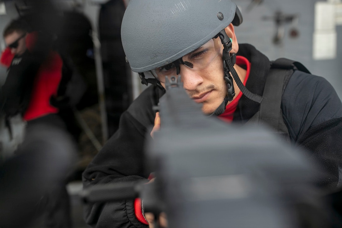 A close-up of a sailor wearing a helmet and protective eyewear while aiming a blurred weapon.