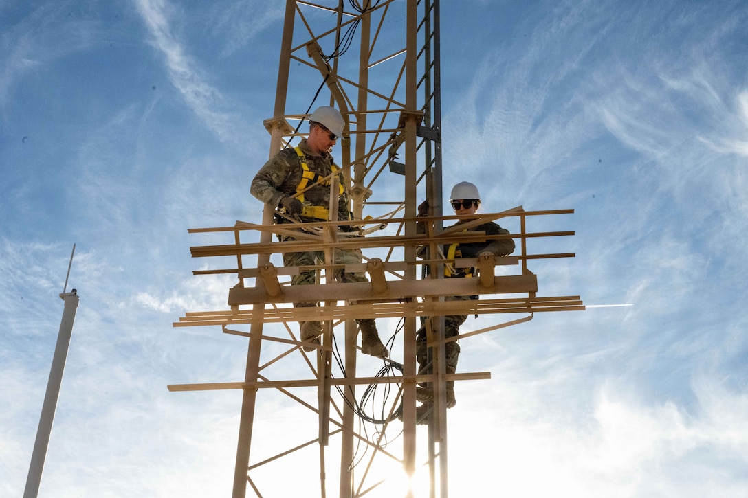 Two airmen wearing protective gear adjust antennae on a tower against a cloudy, blue sky as a bright light beams from the foreground.