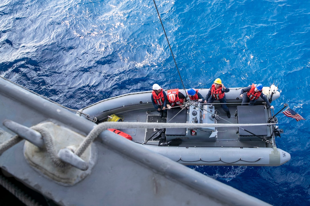 A sailor signals while sitting next to fellow sailors on an inflatable boat as it is lowered into a body of water from a ship as seen from above.