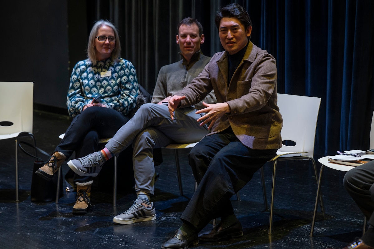 Three individuals sit on chairs in a room with a black floor. One man is wearing a brown jacket and motioning with his hands.