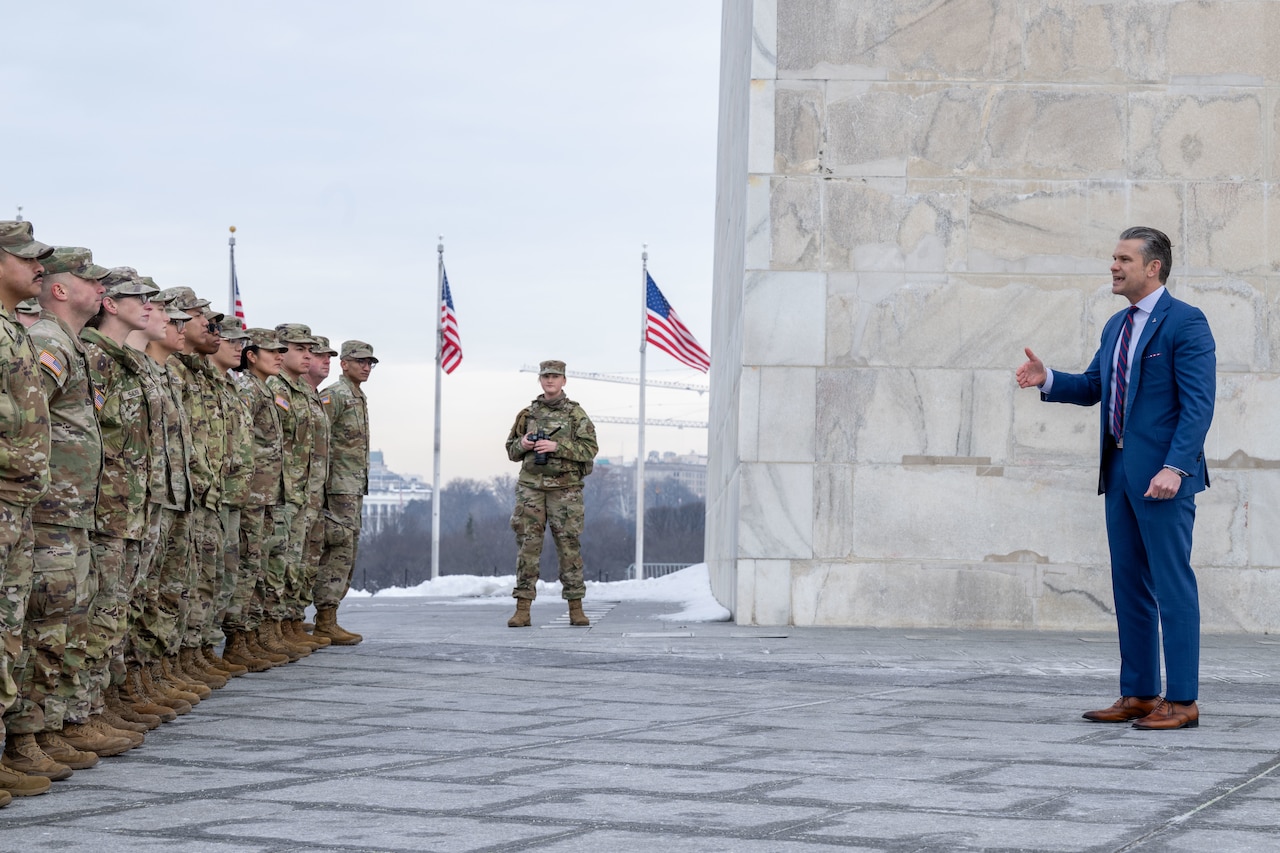 A man in business attire stands near a stone wall outside on a cloudy day, speaking to more than a dozen people wearing camouflage military uniforms; three American flags wave in the background.