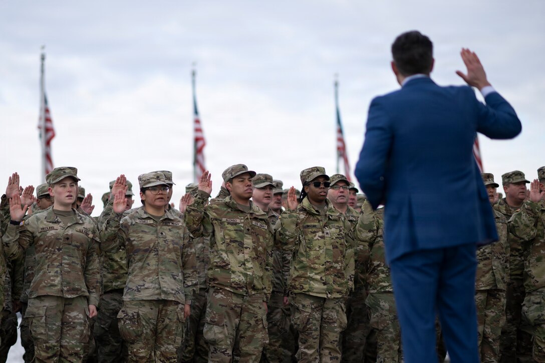 The secretary of war raises his right hand while facing dozens of troops raising their right hands in front of American flags.