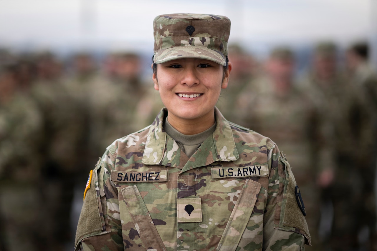 A woman wearing a camouflage military uniform smiles for a portrait; dozens of people in similar attire are blurred in the background.