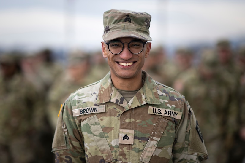 A man wearing a camouflage military uniform smiles for a portrait; dozens of people in similar attire are blurred in the background.