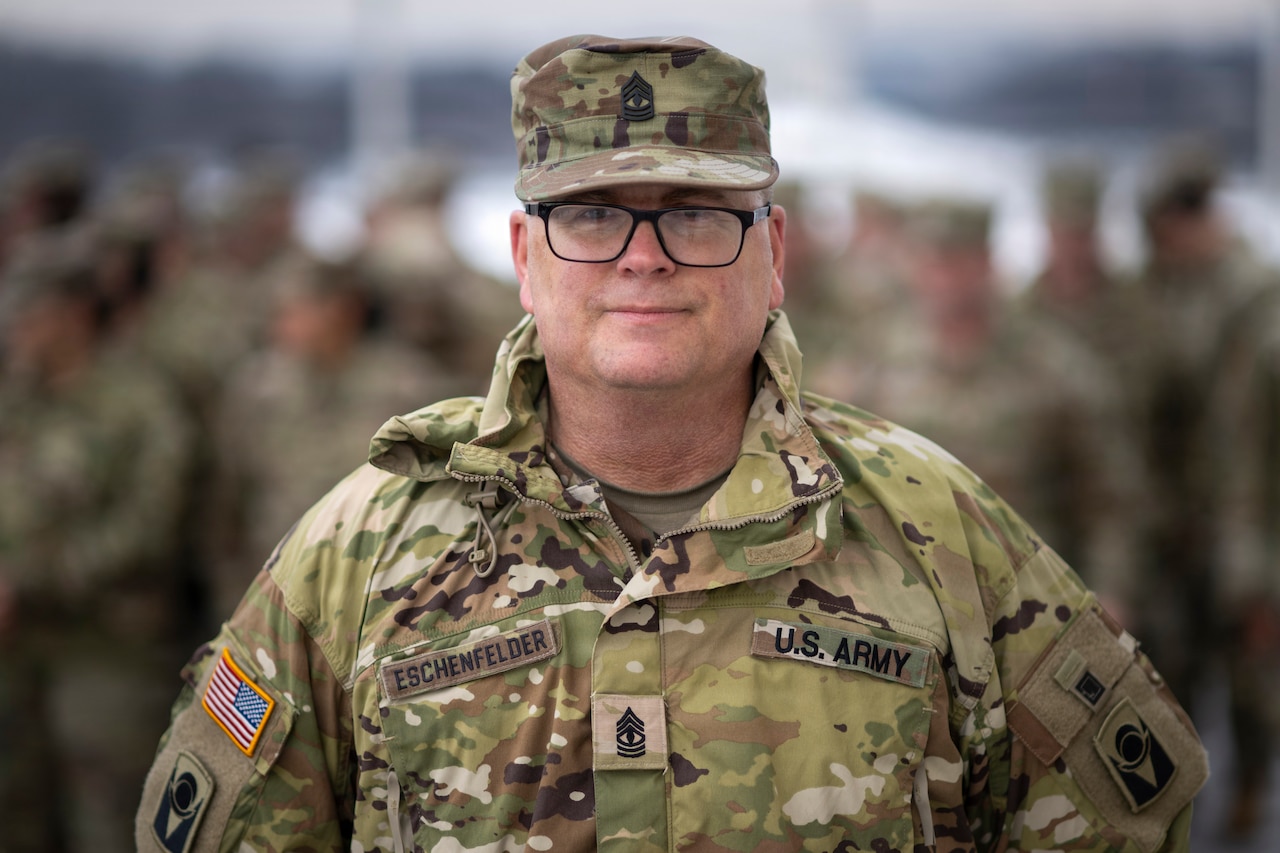 A man wearing a camouflage military uniform smiles for a portrait; dozens of people in similar attire are blurred in the background.