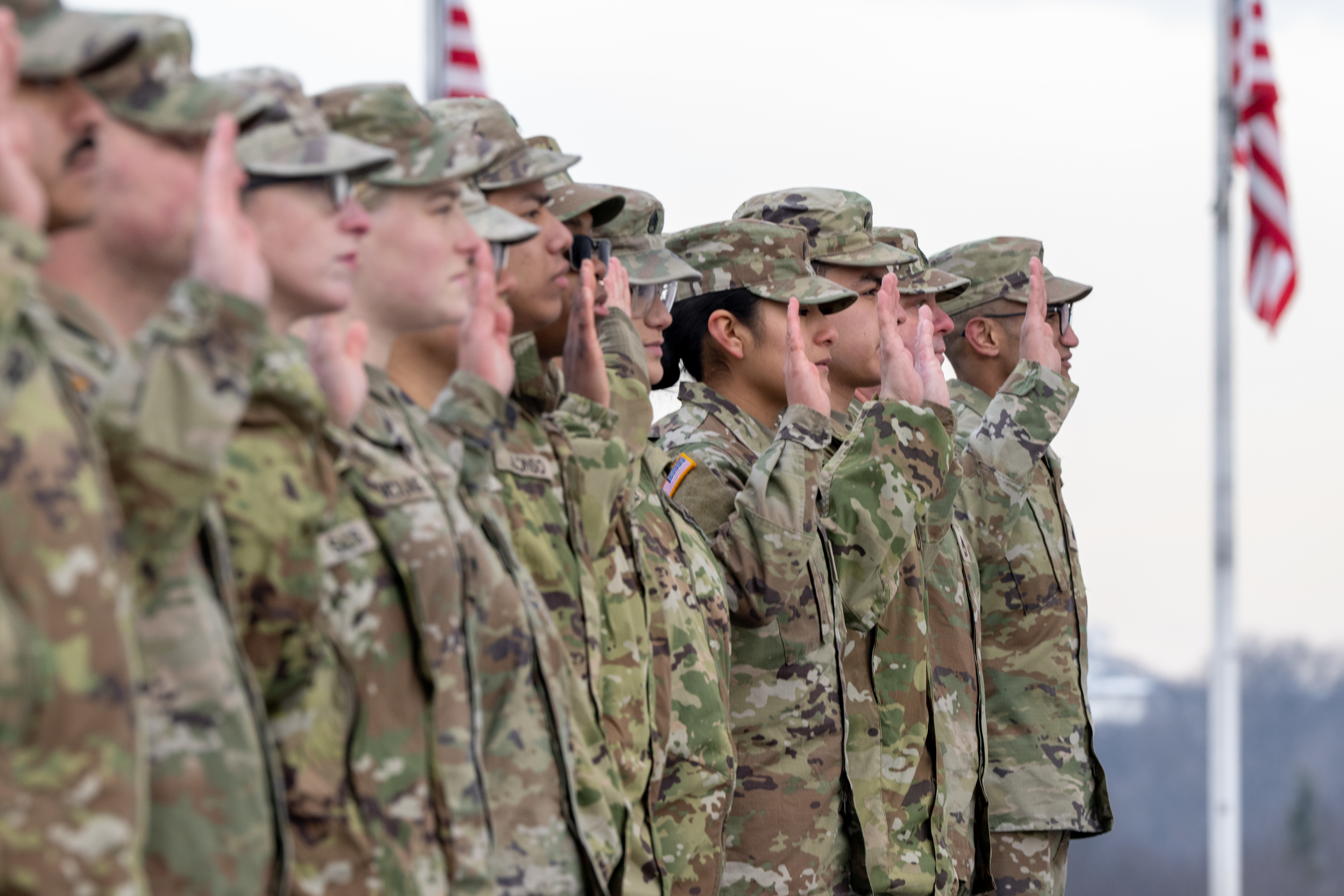About a dozen people wearing camouflage military uniforms stand in formation, raising their right hands; two American flags wave in the background. 