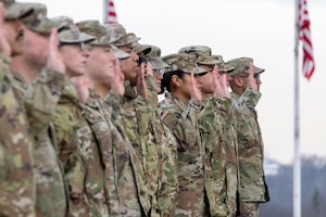 About a dozen people wearing camouflage military uniforms stand in formation, raising their right hands; two American flags wave in the background.