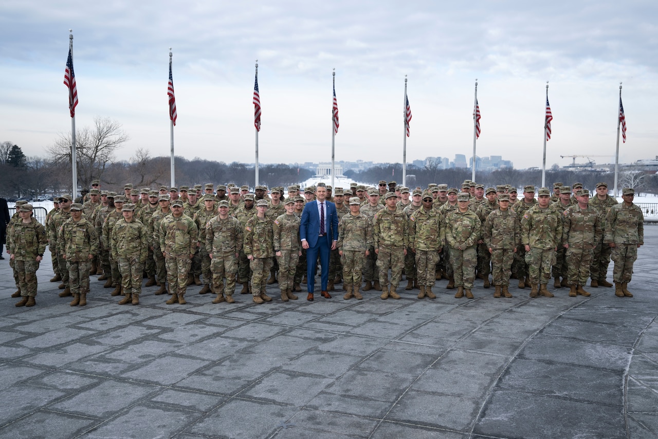 Dozens of people in camouflage military uniforms stand in formation around a man wearing business attire. Behind them stands a row of eight American flags, and in the distance is the Lincoln Memorial.