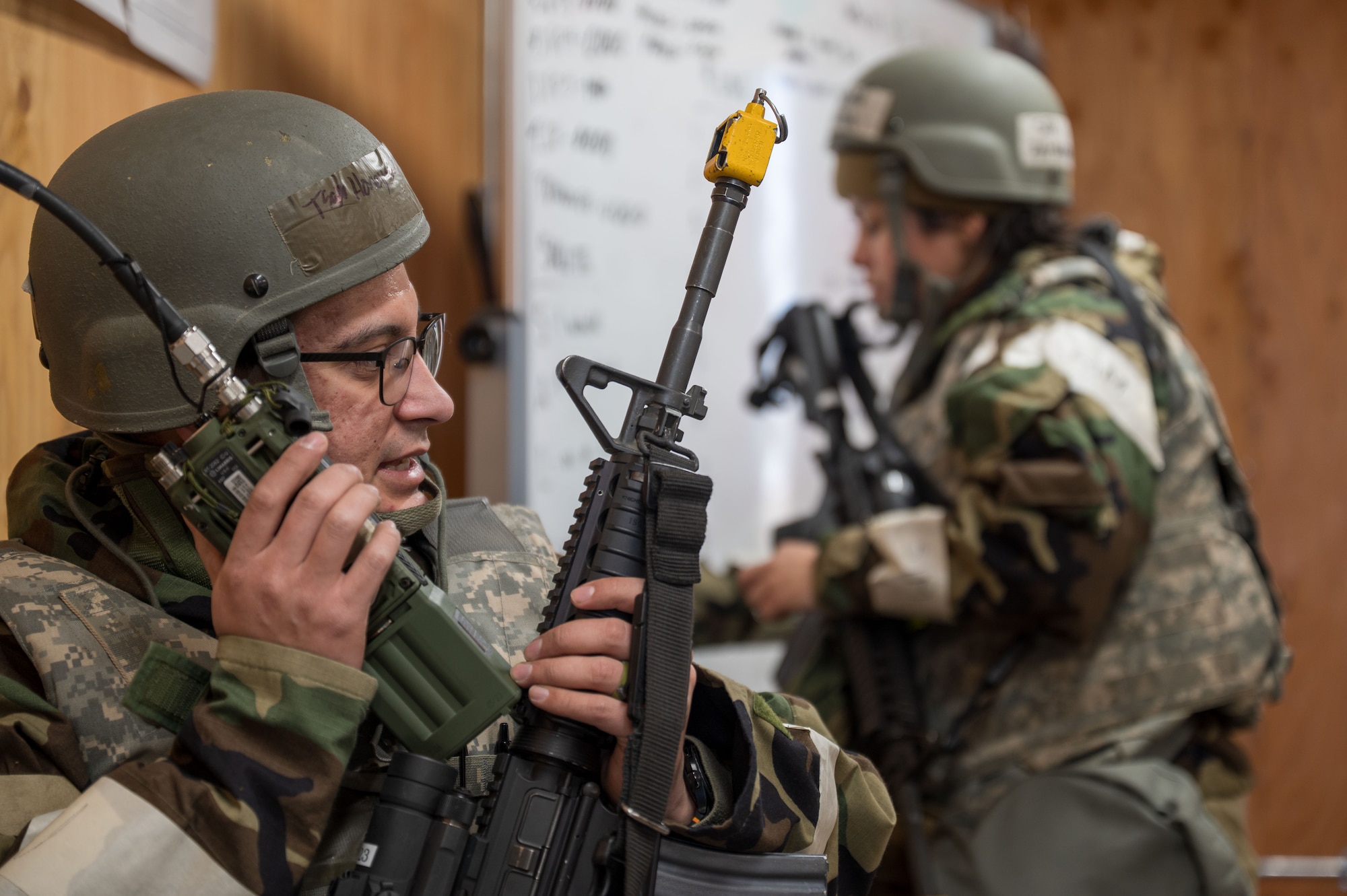 A man speaking into a radio with an assault rifle in hand and a woman in the background writing on a white board.