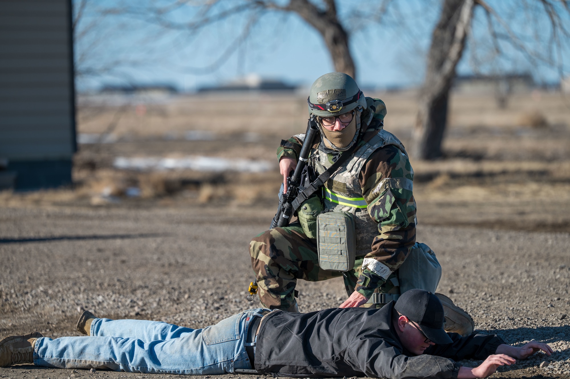 A man in uniform with a helmet and mask kneeling next to a man laying on the ground.