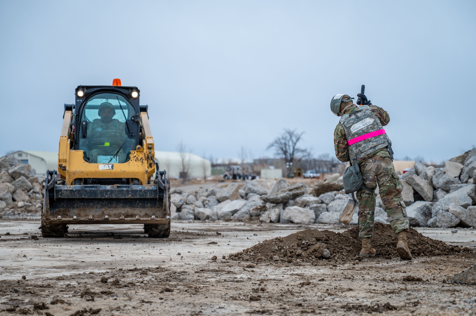 Two men in uniform working at a construction site, one in a skid steer and another shoveling dirt.