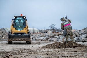 Two men in uniform working at a construction site, one in a skid steer and another shoveling dirt.