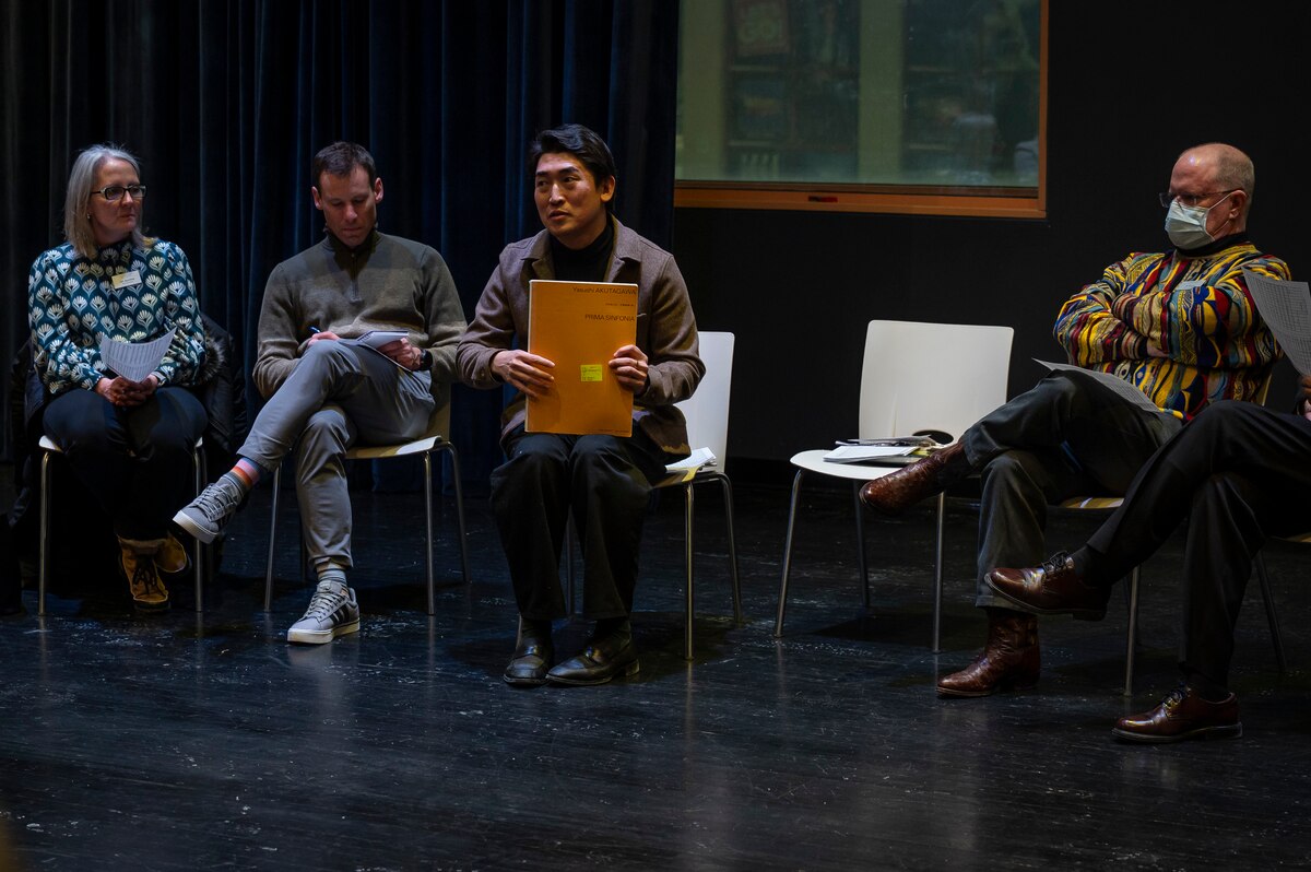 One woman and three men sit in chairs in a room with a black floor. One man holds an orange folder.