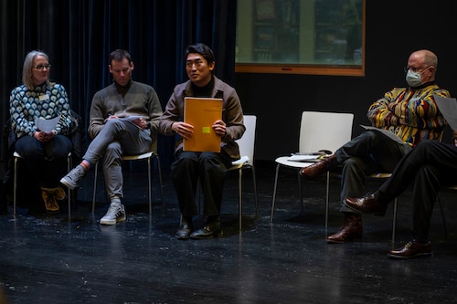 One woman and three men sit in chairs in a room with a black floor. One man holds an orange folder.