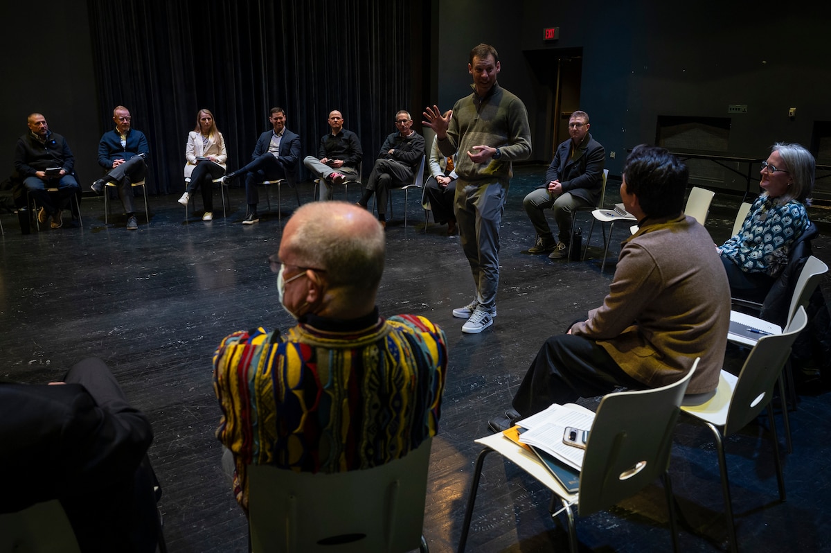Individuals sit in a circle. Man in grey pullover stands in the middle and talks.