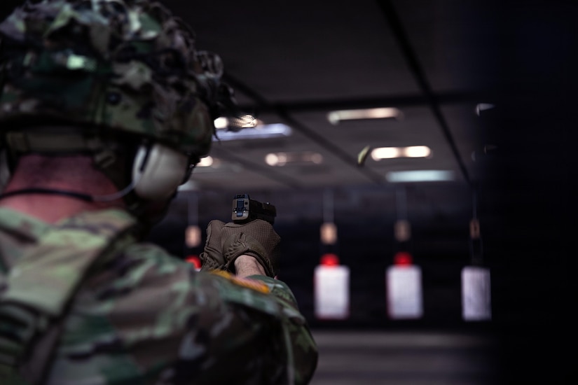 A man in a military camouflage uniform fires a pistol at a target inside a firing range. There is a bullet casing flying away from the gun.