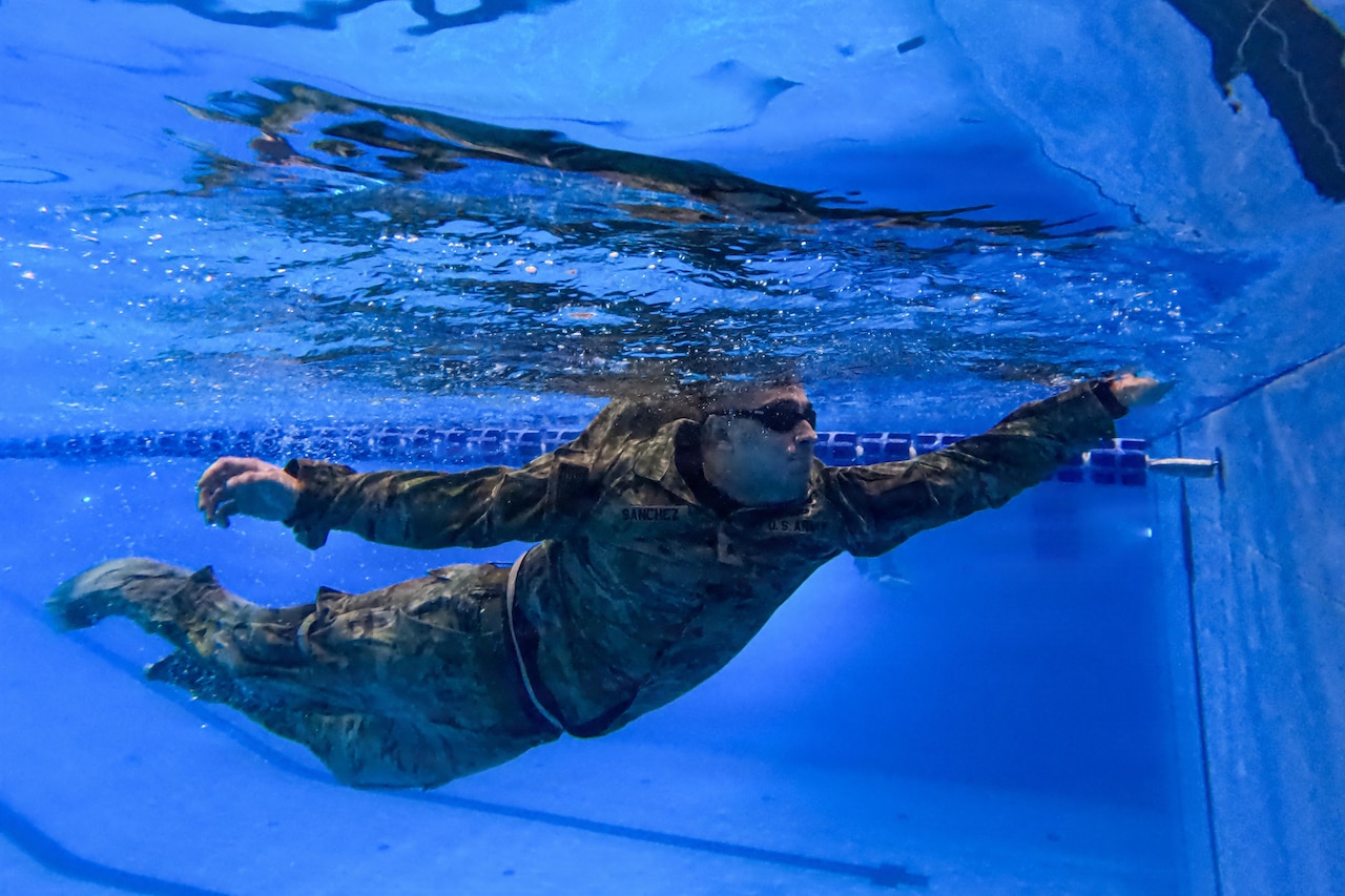 An underwater view of a man in a camouflage military uniform as he reaches his hand out for the pool wall while swimming.