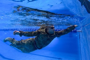 An underwater view of a man in a camouflage military uniform as he reaches his hand out for the pool wall while swimming.