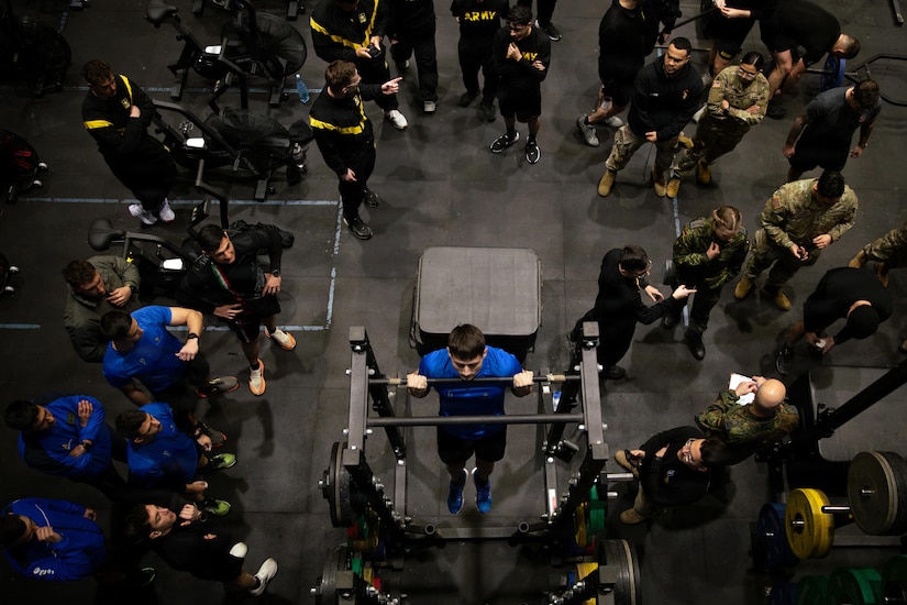 An overhead view of a man in athletic attire holding his chin over a bar on a piece of gym equipment as another man observes him. Two dozen other people in camouflage military uniforms and similar athletic attire are standing around the gym.