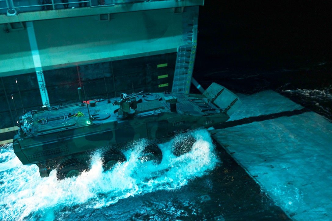 The tires of an amphibious vehicle kick up water as it drives off a ship's well deck in dark conditions.