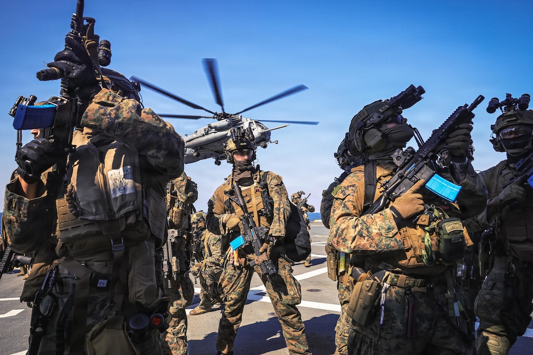 Several people wearing camouflage military uniforms and headgear hold weapons while standing and facing in different directions on a flight deck, as a helicopter hovers in the background.