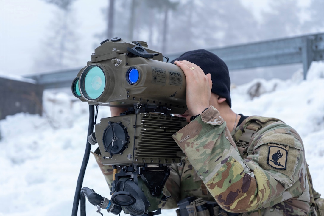 A soldier looks through a lens while standing in a snowy field with trees in the background.
