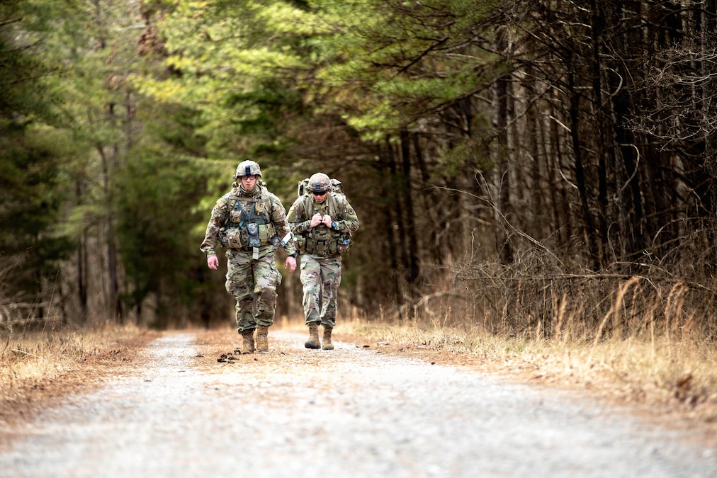 Two soldiers carrying backpacks walk side by side on a paved road through a field.
