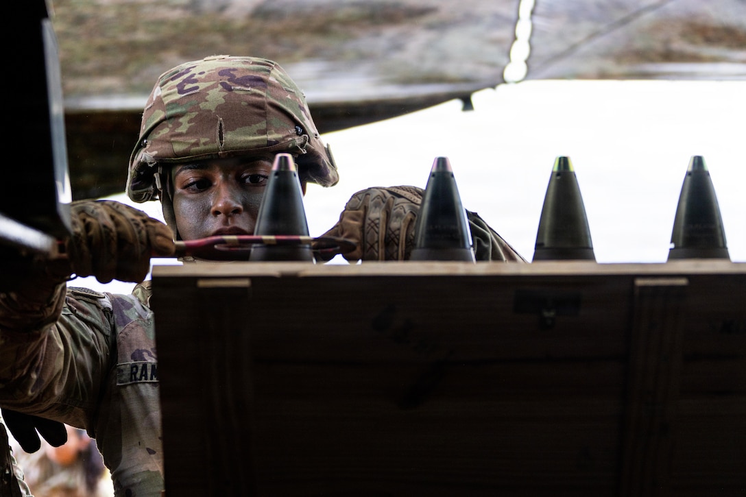 A person wearing a camouflage military uniform, gloves and face paint holds a large tool and works on one of four missiles standing in a row.