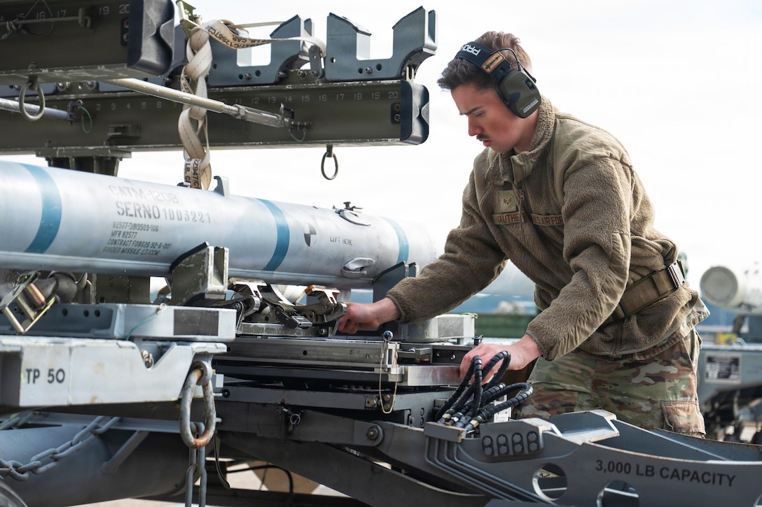 A man wearing a camouflage military uniform looks down and works on a large weapon outside, with a missile on his right.