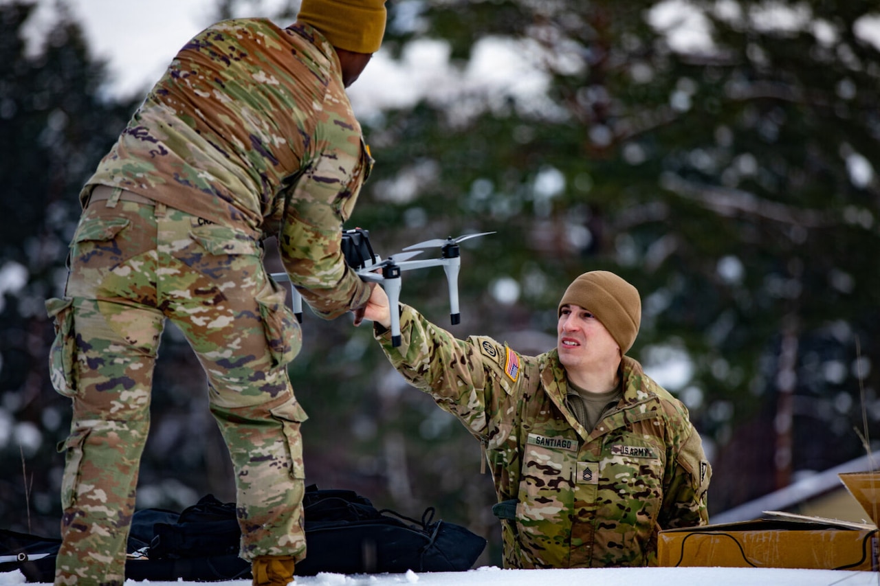 A person wearing a camouflage military uniform hands a drone to a man in similar attire, while standing outside with trees in the background.