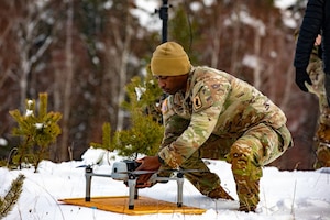 A man wearing a camouflage military uniform places a drone on a wooden board sitting in snow with trees in the background.